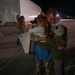 A military service member is reuniting with what appears to be family members, including two children, on an airfield tarmac at night. The emotional embrace shows visible tears on the service member's face, suggesting a homecoming after a deployment. Other uniformed personnel and civilians are visible in the background near a large hangar structure.