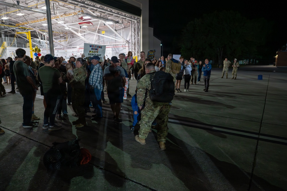 A nighttime military homecoming ceremony taking place on an airfield tarmac outside a large illuminated hangar. Returning service members in camouflage uniforms are being greeted by families and loved ones holding welcome home signs. The scene captures emotional reunions between deployed military personnel and their waiting families.