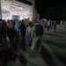 A nighttime military homecoming ceremony taking place on an airfield tarmac outside a large illuminated hangar. Returning service members in camouflage uniforms are being greeted by families and loved ones holding welcome home signs. The scene captures emotional reunions between deployed military personnel and their waiting families.