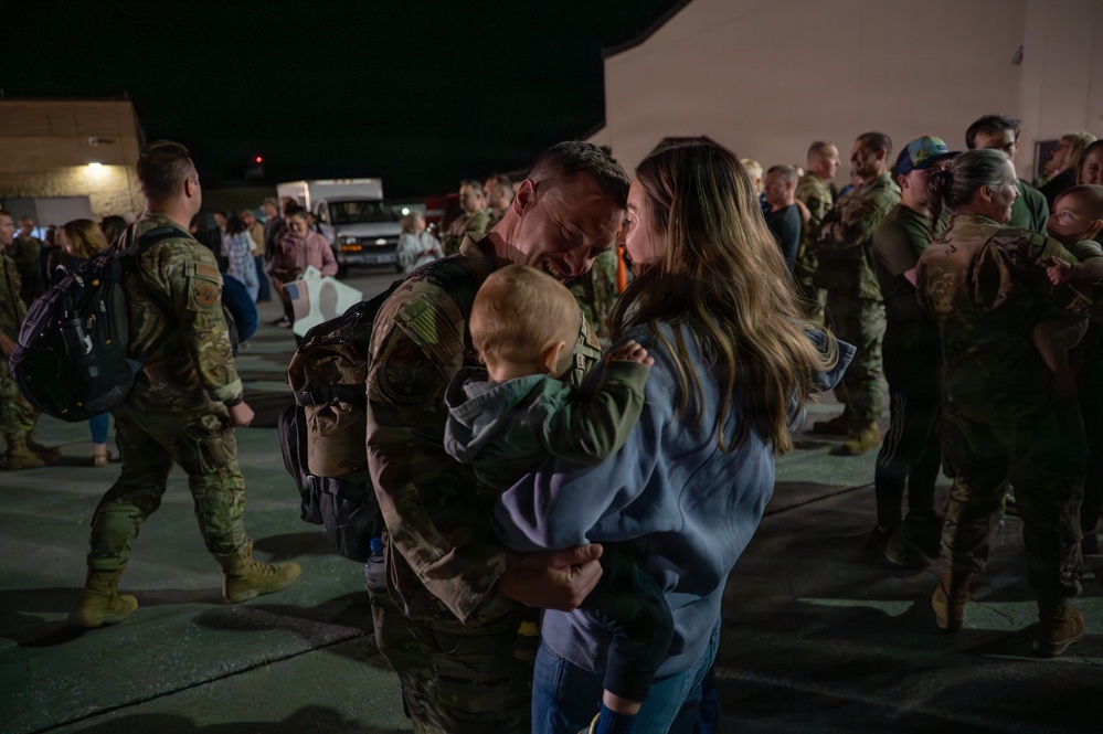 A military service member in OCP (Operational Camouflage Pattern) uniform is reuniting with his family — a woman and a young infant/toddler — in what appears to be a homecoming event on a military installation tarmac at night. The couple is sharing an intimate forehead-to-forehead moment while holding the child between them. In the background, numerous other service members and civilians are engaged in similar reunion interactions.