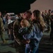 A military service member in OCP (Operational Camouflage Pattern) uniform is reuniting with his family — a woman and a young infant/toddler — in what appears to be a homecoming event on a military installation tarmac at night. The couple is sharing an intimate forehead-to-forehead moment while holding the child between them. In the background, numerous other service members and civilians are engaged in similar reunion interactions.