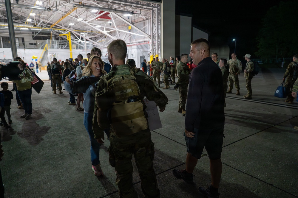 A military homecoming or deployment ceremony is taking place outside a large hangar at night. A soldier with the name tape 'PRATT' visible on their backpack is being greeted by a smiling woman, while a civilian man stands nearby. Multiple uniformed soldiers and civilians are gathered in the background near the illuminated hangar.