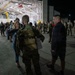 A military homecoming or deployment ceremony is taking place outside a large hangar at night. A soldier with the name tape 'PRATT' visible on their backpack is being greeted by a smiling woman, while a civilian man stands nearby. Multiple uniformed soldiers and civilians are gathered in the background near the illuminated hangar.