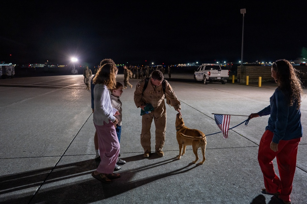 A military service member in tan/khaki flight suit is reuniting with family members on an airfield tarmac at night. The scene appears to be a homecoming event, with the service member bending down to greet a dog held on a leash by a woman holding a small American flag. Additional personnel and vehicles are visible in the background.