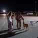 A military service member in tan/khaki flight suit is reuniting with family members on an airfield tarmac at night. The scene appears to be a homecoming event, with the service member bending down to greet a dog held on a leash by a woman holding a small American flag. Additional personnel and vehicles are visible in the background.