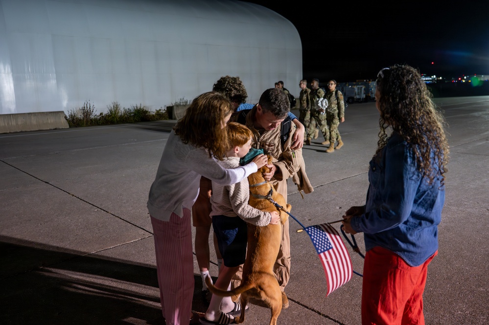 A nighttime military homecoming scene on what appears to be an airfield tarmac. A service member in a flight suit is being greeted by family members including a woman, a young child, and a dog, while another person holding American flags stands nearby. Additional uniformed personnel can be seen walking in the background.