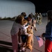 A nighttime military homecoming scene on what appears to be an airfield tarmac. A service member in a flight suit is being greeted by family members including a woman, a young child, and a dog, while another person holding American flags stands nearby. Additional uniformed personnel can be seen walking in the background.