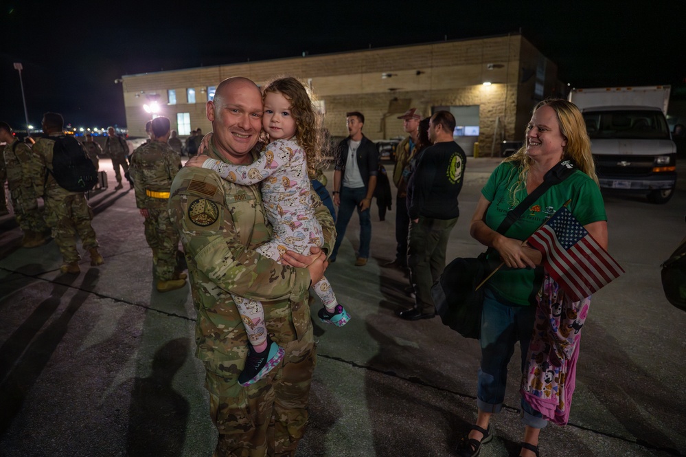 A military service member in MultiCam/OCP camouflage uniform is reuniting with family at what appears to be a homecoming event on a military installation at night. He is holding a young child while a woman in a green shirt holds a small American flag and smiles nearby. Other uniformed personnel and civilians are visible in the background near a building.