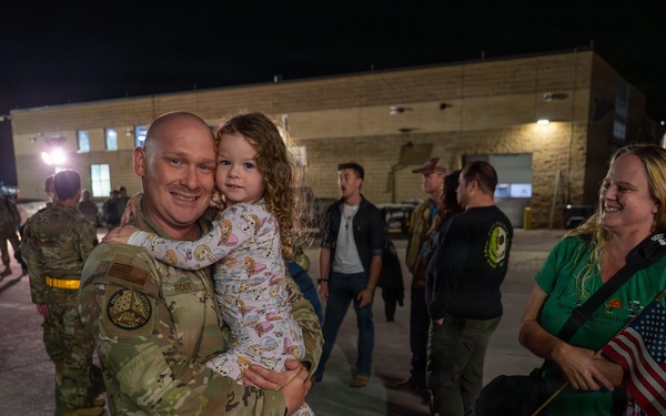 A military service member in MultiCam/OCP camouflage uniform is reuniting with family at what appears to be a homecoming event on a military installation at night. He is holding a young child while a woman in a green shirt holds a small American flag and smiles nearby. Other uniformed personnel and civilians are visible in the background near a building.