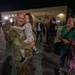 A military service member in MultiCam/OCP camouflage uniform is reuniting with family at what appears to be a homecoming event on a military installation at night. He is holding a young child while a woman in a green shirt holds a small American flag and smiles nearby. Other uniformed personnel and civilians are visible in the background near a building.