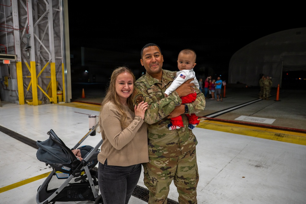 A military service member in OCP (Operational Camouflage Pattern) uniform is reuniting with his family on what appears to be an airfield tarmac at night. He is holding a young infant dressed in patriotic red, white, and blue clothing while standing beside a smiling woman who is holding a baby stroller. The setting and context suggest a military homecoming event.