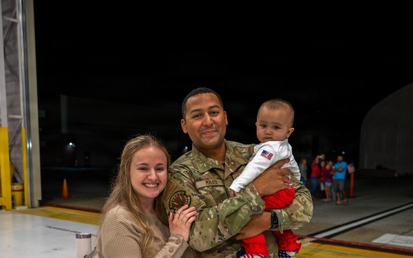 A military service member in OCP (Operational Camouflage Pattern) uniform is reuniting with his family on what appears to be an airfield tarmac at night. He is holding a young infant dressed in patriotic red, white, and blue clothing while standing beside a smiling woman who is holding a baby stroller. The setting and context suggest a military homecoming event.