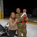 A military service member in OCP (Operational Camouflage Pattern) uniform is reuniting with his family on what appears to be an airfield tarmac at night. He is holding a young infant dressed in patriotic red, white, and blue clothing while standing beside a smiling woman who is holding a baby stroller. The setting and context suggest a military homecoming event.