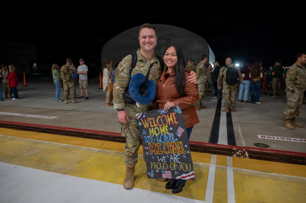 A military homecoming scene at night on what appears to be an airfield or tarmac. A soldier in OCP (Operational Camouflage Pattern) uniform is posing with a woman holding a handmade welcome home sign addressed to 'Michael.' Other military personnel and civilians are visible in the background, suggesting a larger unit return event.