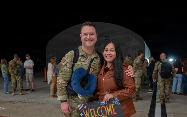 A military homecoming scene at night on what appears to be an airfield or tarmac. A soldier in OCP (Operational Camouflage Pattern) uniform is posing with a woman holding a handmade welcome home sign addressed to 'Michael.' Other military personnel and civilians are visible in the background, suggesting a larger unit return event.
