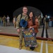 A military homecoming scene at night on what appears to be an airfield or tarmac. A soldier in OCP (Operational Camouflage Pattern) uniform is posing with a woman holding a handmade welcome home sign addressed to 'Michael.' Other military personnel and civilians are visible in the background, suggesting a larger unit return event.