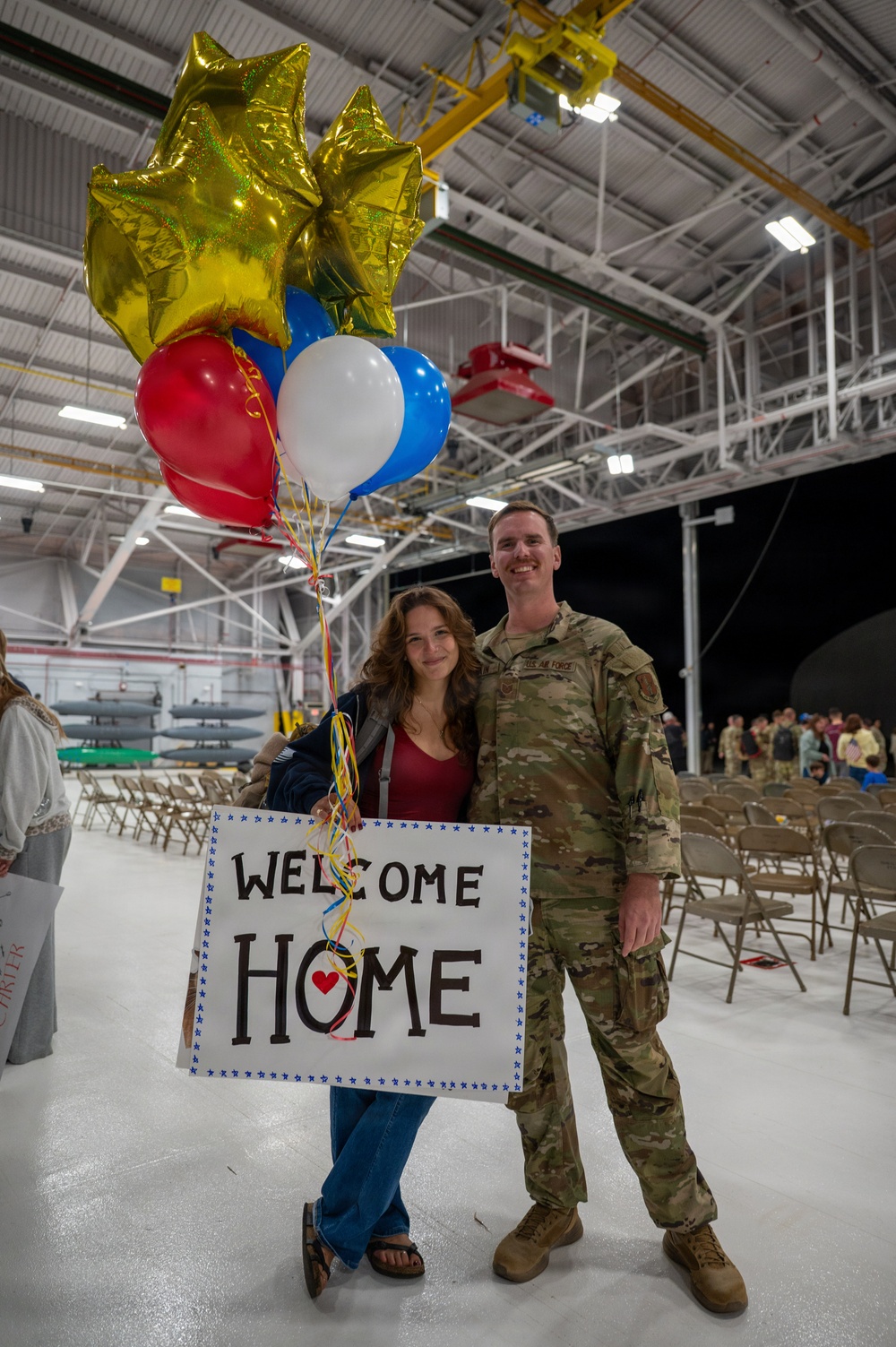 A military homecoming ceremony taking place inside an aircraft hangar. A U.S. Air Force service member in OCP (Operational Camouflage Pattern) uniform poses with a civilian woman holding red, white, and blue balloons along with gold star mylar balloons and a handmade 'Welcome Home' sign. The setting includes rows of folding chairs and what appear to be munitions or aircraft components visible in the background.