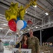 A military homecoming ceremony taking place inside an aircraft hangar. A U.S. Air Force service member in OCP (Operational Camouflage Pattern) uniform poses with a civilian woman holding red, white, and blue balloons along with gold star mylar balloons and a handmade 'Welcome Home' sign. The setting includes rows of folding chairs and what appear to be munitions or aircraft components visible in the background.