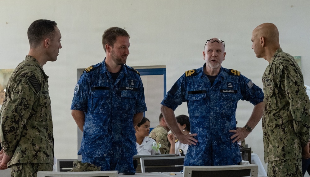 Four military personnel are engaged in conversation in what appears to be a meeting or dining facility. Two individuals wear US Navy digital camouflage uniforms (NWU Type III), while two others wear Belgian Navy blue digital camouflage uniforms with gold shoulder rank insignia. The setting suggests a multinational military meeting or liaison event.