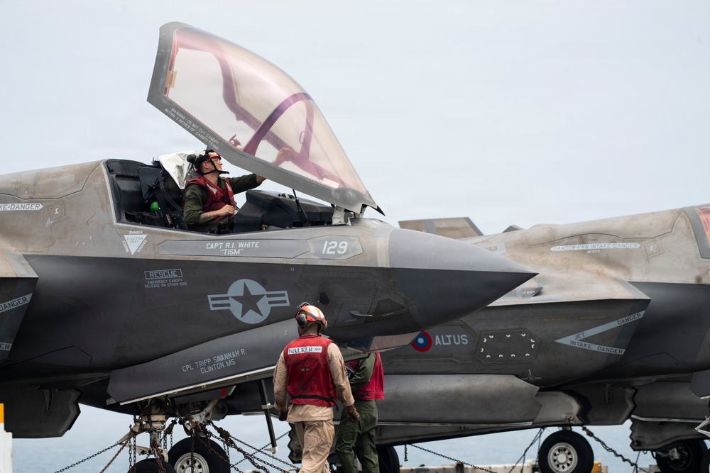 A military jet aircraft, identified as aircraft number 129, is shown on what appears to be the deck of an aircraft carrier. A pilot is seated in the open cockpit while ground crew personnel in red vests attend to the aircraft. The scene depicts pre-flight or post-flight operations on a naval vessel.
