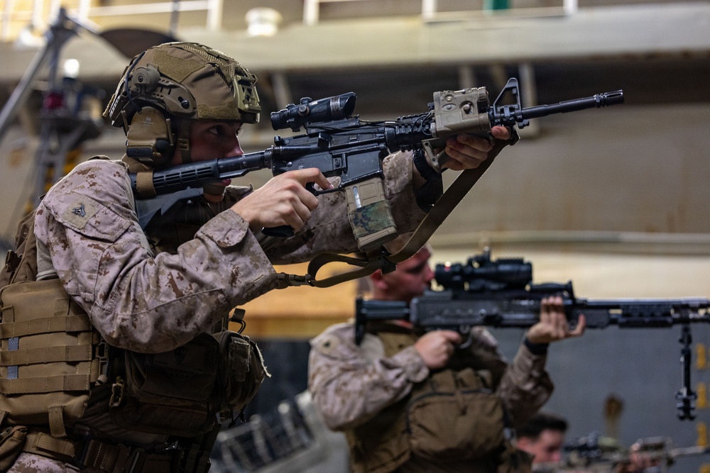 Two military personnel in MARPAT desert camouflage uniforms are shown in a tactical aiming stance, holding rifles in what appears to be an indoor military facility or ship hangar deck. The foreground soldier is equipped with full combat gear including a tactical helmet, plate carrier, and an M4-style carbine with optics and accessories. The image captures what appears to be a training exercise or readiness drill.