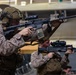 Two military personnel in MARPAT desert camouflage uniforms are shown in a tactical aiming stance, holding rifles in what appears to be an indoor military facility or ship hangar deck. The foreground soldier is equipped with full combat gear including a tactical helmet, plate carrier, and an M4-style carbine with optics and accessories. The image captures what appears to be a training exercise or readiness drill.