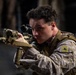 A male service member in MARPAT (Marine pattern) camouflage uniform is aiming a rifle equipped with an optical scope and tactical accessories. He is wearing a rank insignia patch on his left sleeve, consistent with a lower enlisted Marine rank. The image is a close-up, well-lit photograph taken in a low-light environment.