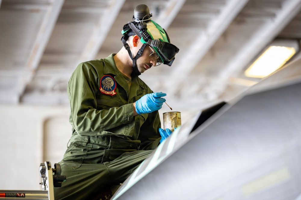 A military aviation technician in olive green coveralls is performing maintenance work on what appears to be an aircraft surface, likely inside a hangar. The individual is using a brush and a small container, suggesting painting, coating, or sealant application on the aircraft. Protective equipment including safety glasses, blue nitrile gloves, and a helmet-mounted device are visible.