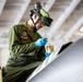 A military aviation technician in olive green coveralls is performing maintenance work on what appears to be an aircraft surface, likely inside a hangar. The individual is using a brush and a small container, suggesting painting, coating, or sealant application on the aircraft. Protective equipment including safety glasses, blue nitrile gloves, and a helmet-mounted device are visible.