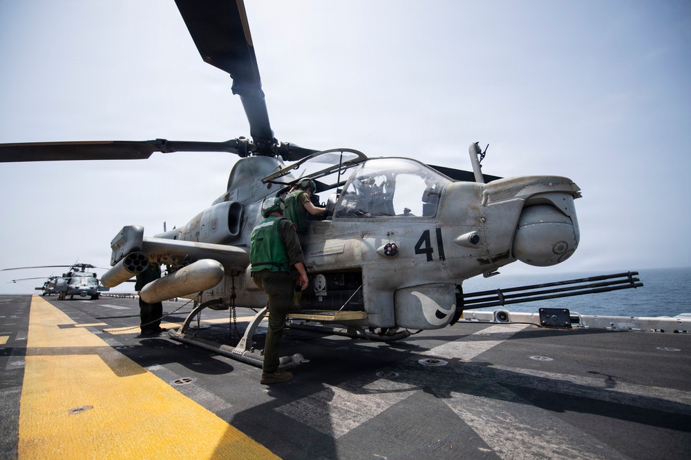 A military attack helicopter marked with the number '41' is positioned on the flight deck of a naval vessel at sea. Ground crew personnel in green vests are conducting pre-flight or maintenance checks on the aircraft. Additional helicopters are visible in the background on the same flight deck.