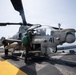 A military attack helicopter marked with the number '41' is positioned on the flight deck of a naval vessel at sea. Ground crew personnel in green vests are conducting pre-flight or maintenance checks on the aircraft. Additional helicopters are visible in the background on the same flight deck.