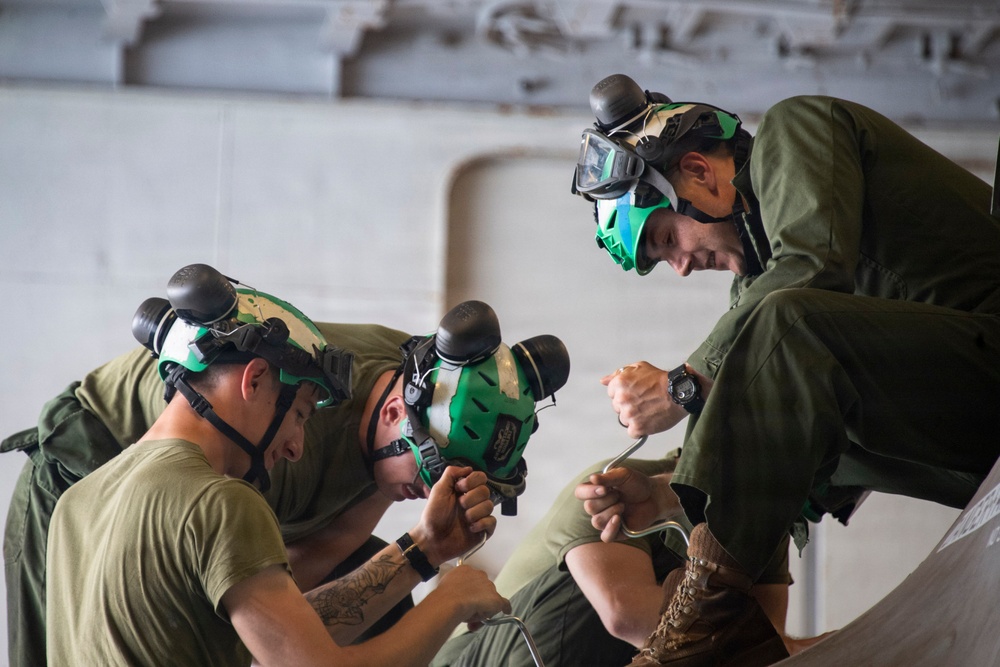 Three individuals wearing military-style olive drab uniforms and green protective helmets with goggles are gathered closely together, appearing to work on or examine something at a lower surface. The person on the right is positioned higher and leaning in to observe, while the other two are bent over in focused concentration. The setting appears to be an indoor stone or concrete structure.