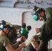 Three individuals wearing military-style olive drab uniforms and green protective helmets with goggles are gathered closely together, appearing to work on or examine something at a lower surface. The person on the right is positioned higher and leaning in to observe, while the other two are bent over in focused concentration. The setting appears to be an indoor stone or concrete structure.