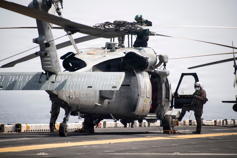 A military helicopter, consistent with an MH-60 Seahawk variant, is positioned on the flight deck of a naval vessel at sea. At least two personnel in flight gear and helmets are conducting maintenance or pre-flight inspection activities. The overcast sky and visible ocean horizon confirm an open-water shipboard environment.