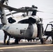 A military helicopter, consistent with an MH-60 Seahawk variant, is positioned on the flight deck of a naval vessel at sea. At least two personnel in flight gear and helmets are conducting maintenance or pre-flight inspection activities. The overcast sky and visible ocean horizon confirm an open-water shipboard environment.