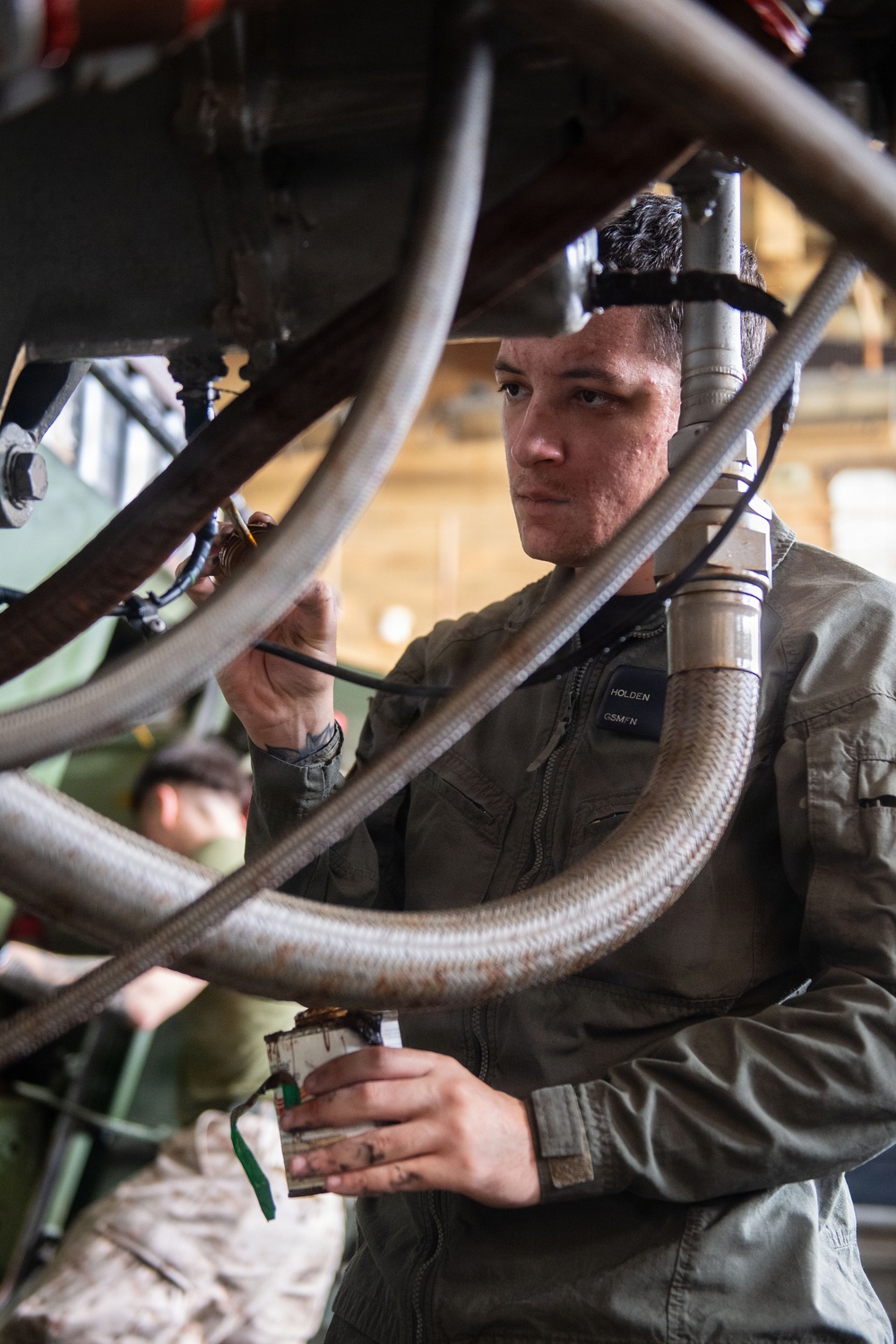 A military technician identified by a name tape reading 'HOLDEN' and a rating/designation patch reading 'GSMFN' is performing maintenance work on mechanical equipment surrounded by hoses and cables. He is wearing an olive/sage green coverall uniform and is focused on working with his hands on a component within a complex assembly. A second individual in military uniform is partially visible in the background.