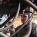 A military technician identified by a name tape reading 'HOLDEN' and a rating/designation patch reading 'GSMFN' is performing maintenance work on mechanical equipment surrounded by hoses and cables. He is wearing an olive/sage green coverall uniform and is focused on working with his hands on a component within a complex assembly. A second individual in military uniform is partially visible in the background.