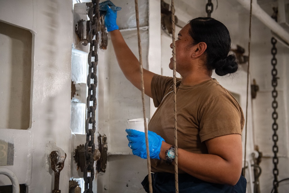 A person wearing a tan/olive military-style t-shirt and blue nitrile gloves is performing maintenance work, appearing to paint or clean a white metal surface aboard what appears to be a naval vessel. They are holding a small white container (likely paint or cleaning solution) in one hand while applying material to the surface with the other. The environment features heavy chains, ropes, and metal hardware consistent with a ship's interior or deck area.