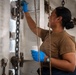 A person wearing a tan/olive military-style t-shirt and blue nitrile gloves is performing maintenance work, appearing to paint or clean a white metal surface aboard what appears to be a naval vessel. They are holding a small white container (likely paint or cleaning solution) in one hand while applying material to the surface with the other. The environment features heavy chains, ropes, and metal hardware consistent with a ship's interior or deck area.