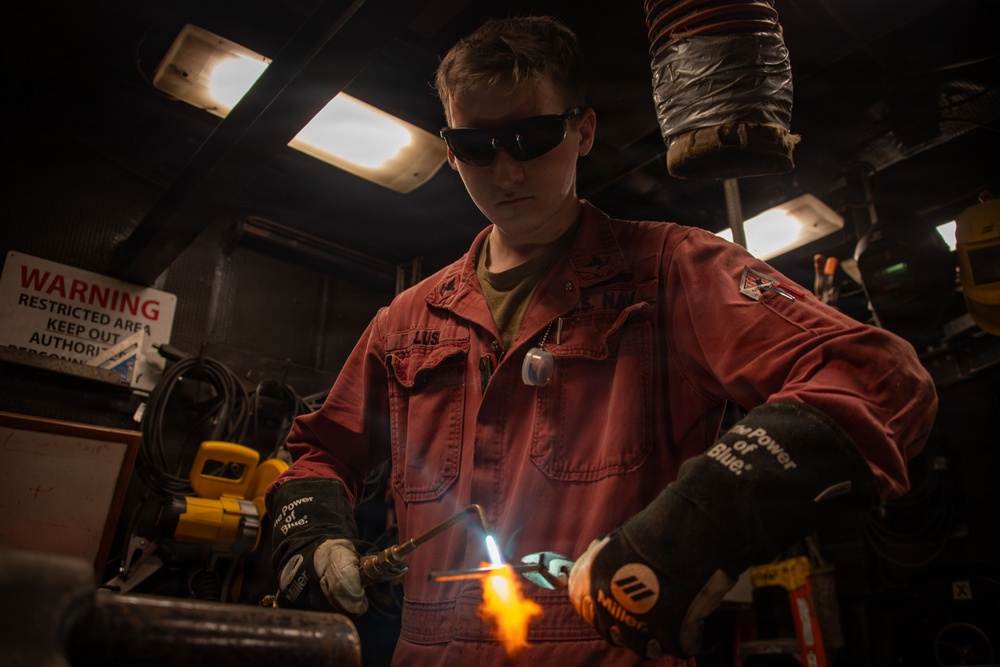 A young worker in red coveralls is performing welding or torch-cutting work in an industrial shop environment. They are wearing protective dark-tinted safety glasses and Miller-branded welding gloves while operating a torch that produces visible orange and blue flames. The setting appears to be a restricted military or industrial maintenance facility.