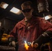 A young worker in red coveralls is performing welding or torch-cutting work in an industrial shop environment. They are wearing protective dark-tinted safety glasses and Miller-branded welding gloves while operating a torch that produces visible orange and blue flames. The setting appears to be a restricted military or industrial maintenance facility.