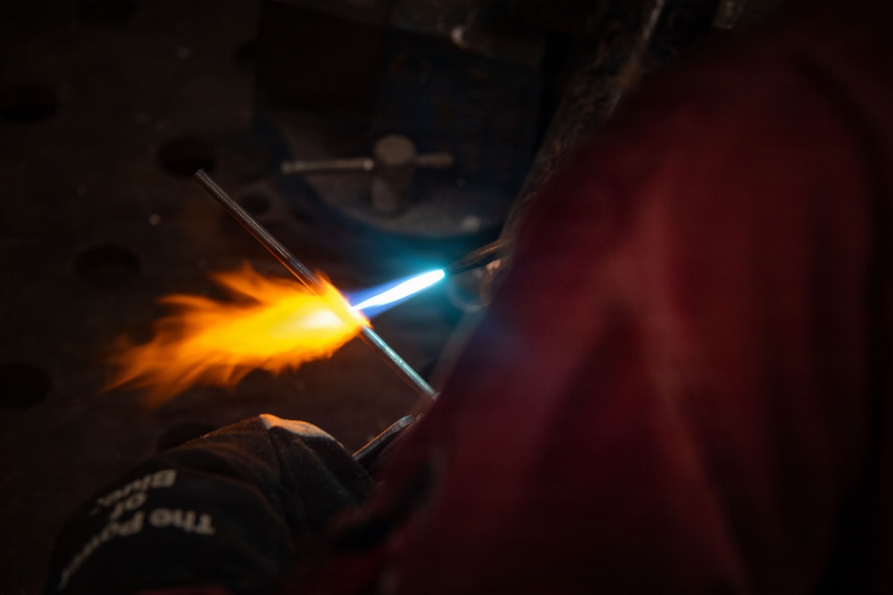 A person wearing a protective glove is using a torch to heat a glass rod or metal rod, producing a vivid orange and yellow flame with a bright blue torch flame visible. The scene is set in a dark workshop or studio environment, suggesting flameworking or metalworking activity. The dramatic lighting from the flame contrasts sharply against the dark background.