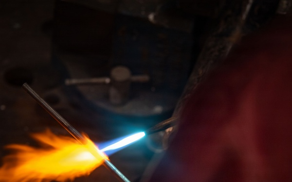 A person wearing a protective glove is using a torch to heat a glass rod or metal rod, producing a vivid orange and yellow flame with a bright blue torch flame visible. The scene is set in a dark workshop or studio environment, suggesting flameworking or metalworking activity. The dramatic lighting from the flame contrasts sharply against the dark background.