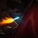A person wearing a protective glove is using a torch to heat a glass rod or metal rod, producing a vivid orange and yellow flame with a bright blue torch flame visible. The scene is set in a dark workshop or studio environment, suggesting flameworking or metalworking activity. The dramatic lighting from the flame contrasts sharply against the dark background.