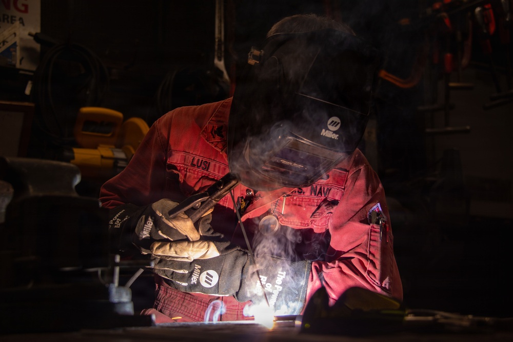 A welder is actively welding metal in a dark workshop environment, with visible sparks, arc light, and smoke rising from the work area. The individual is wearing full protective gear including a Miller brand welding helmet and Miller brand gloves, along with a red work uniform bearing text that includes 'NAVY'. The dramatic low-key lighting highlights the welding arc and smoke against the dark background.