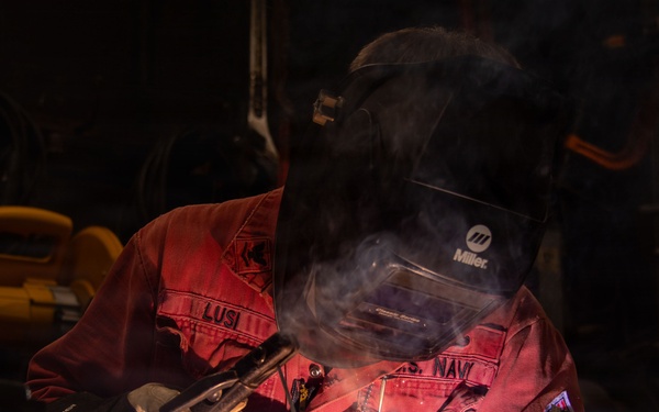 A welder is actively welding metal in a dark workshop environment, with visible sparks, arc light, and smoke rising from the work area. The individual is wearing full protective gear including a Miller brand welding helmet and Miller brand gloves, along with a red work uniform bearing text that includes 'NAVY'. The dramatic low-key lighting highlights the welding arc and smoke against the dark background.