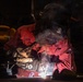 A welder is actively welding metal in a dark workshop environment, with visible sparks, arc light, and smoke rising from the work area. The individual is wearing full protective gear including a Miller brand welding helmet and Miller brand gloves, along with a red work uniform bearing text that includes 'NAVY'. The dramatic low-key lighting highlights the welding arc and smoke against the dark background.