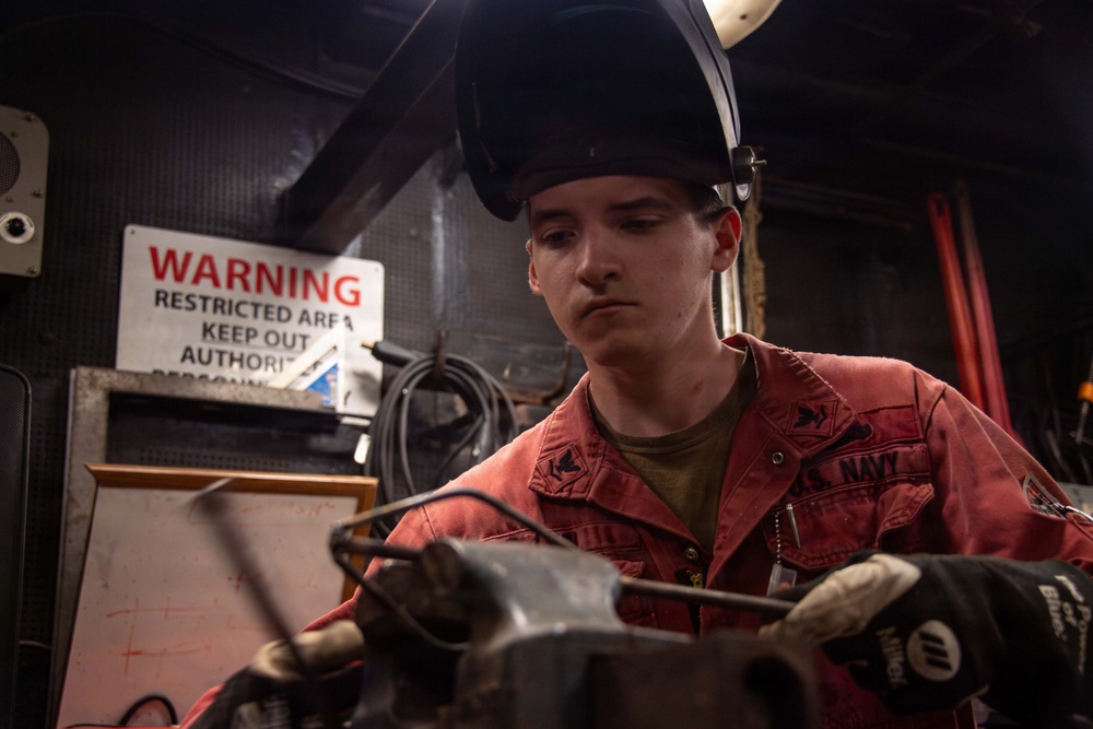 A U.S. Navy sailor wearing a red coverall uniform and a welding helmet (raised) is working with equipment in what appears to be a shipboard workshop or machinery space. The individual is wearing protective gloves and appears to be operating or adjusting metalworking equipment. A warning sign indicating a restricted area is visible on the wall behind them.