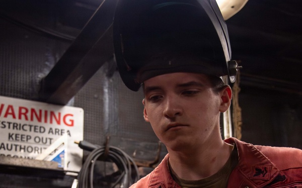 A U.S. Navy sailor wearing a red coverall uniform and a welding helmet (raised) is working with equipment in what appears to be a shipboard workshop or machinery space. The individual is wearing protective gloves and appears to be operating or adjusting metalworking equipment. A warning sign indicating a restricted area is visible on the wall behind them.
