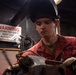 A U.S. Navy sailor wearing a red coverall uniform and a welding helmet (raised) is working with equipment in what appears to be a shipboard workshop or machinery space. The individual is wearing protective gloves and appears to be operating or adjusting metalworking equipment. A warning sign indicating a restricted area is visible on the wall behind them.